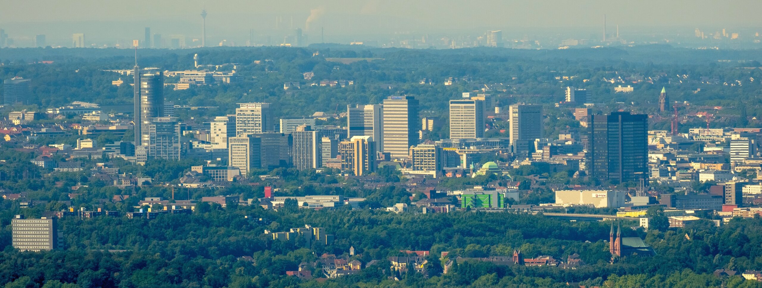 Schauen Sie mal ganz genau hin. Auf diesem Luftbild hat Hans Blossey bei außergewöhnlich klarer Sicht mit einem Teleobjektiv die Skyline von gleich zwei Städten eingefangen: Vorne im Bild sind das Essener Hochhausviertel mit RWE-Turm (links), neuer Schenker-Zentrale und Rheinstahl-Haus (rechts) sowie das Essener Rathaus (vorne, rechts außen) zu sehen. Am Horizont grüßt obendrein der Düsseldorfer Rheinturm. Der Landtag direkt daneben ist zwar nicht in Sicht, aber das Dreischeibenhaus (ehemaligen Thyssen-Krupp-Zentrale, etwas weiter links) ist ebenso deutlich zu erkennen wie das Stadttor (Sitz der Staatskanzlei) und der Victoria-Tower (Sitz der Ergo-Versicherung). Rechts im Bild ist oben neben einer Rauchwolke sogar eine Rheinquerung zu sehen: die Theodor-Heuss-Brücke. Das Flugzeug mit Hans Blossey war in diesem Moment übrigens über Gelsenkirchen (südlich des Zoom-Zoos) unterwegs. Luftlinie von dort bis zum Essener Rathaus: etwa 11,5 Kilometer. Die direkte Distanz bis nach Düsseldorf: etwa 40 Kilometer. Noch zwei Orientierungspunkte im Essener Bildvordergrund: Hinter dem Grüngürtel ragt halblinks der Turm der Feuerwehrhauptwache an der Eisernen Hand empor, vorne rechts im Bild der Doppelturm der Kirche St. Nikolaus in Stoppenberg.