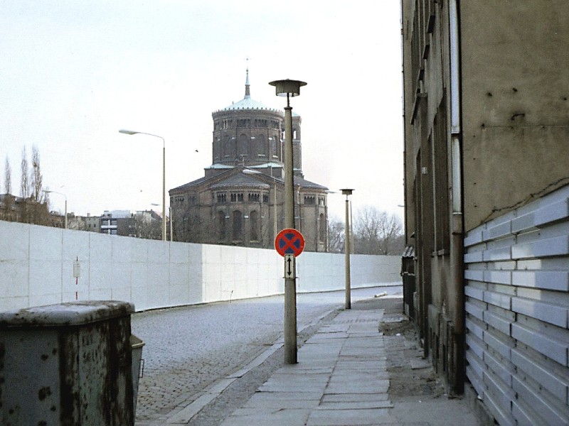 Die weiß gestrichene Hinterlandmauer markierte den Beginn des Grenzstreifens von der Ostseite her, während die Thomaskirche im Hintergrund bereits im Westen steht. Die Überwachung begann jedoch weit vor dieser Mauer: Mit der sogenannten „Hinterlandsicherung“ kontrollierten Stasi, Grenztruppen und Polizei das Gebiet nahe dem Grenzstreifen. Für den Aufenthalt war ein Berechtigungsschein erforderlich.