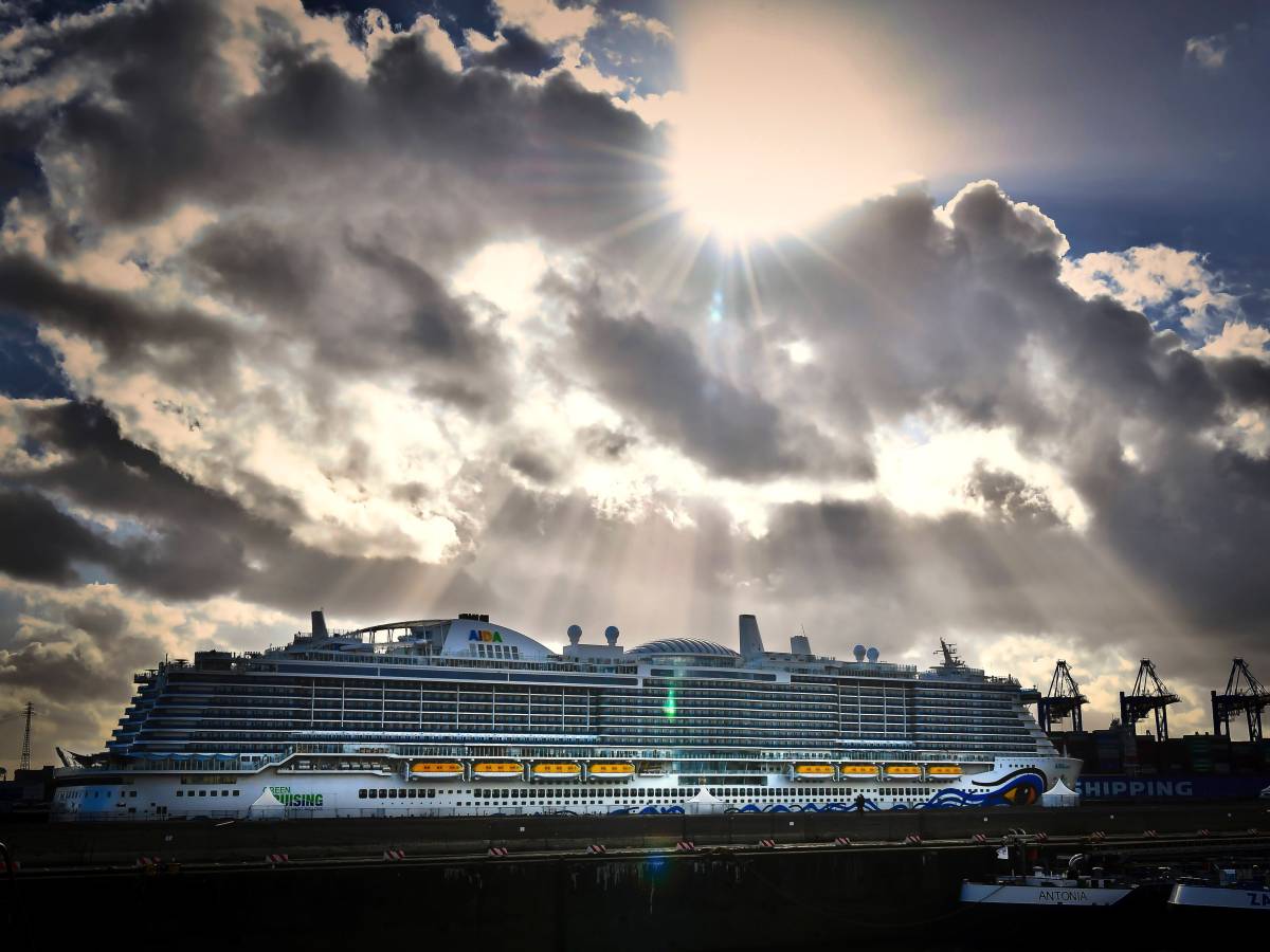 Ein Kreuzfahrt-Schiff der Aida. Über ihm türmen sich Wolken, durch die die Sonne bricht.