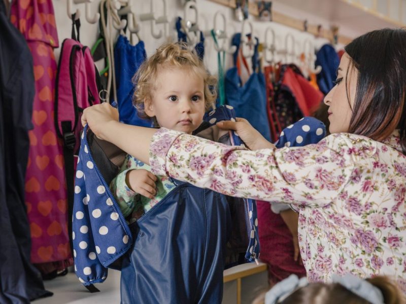 Kita-Erzieherin zieht Kind eine Jacke an