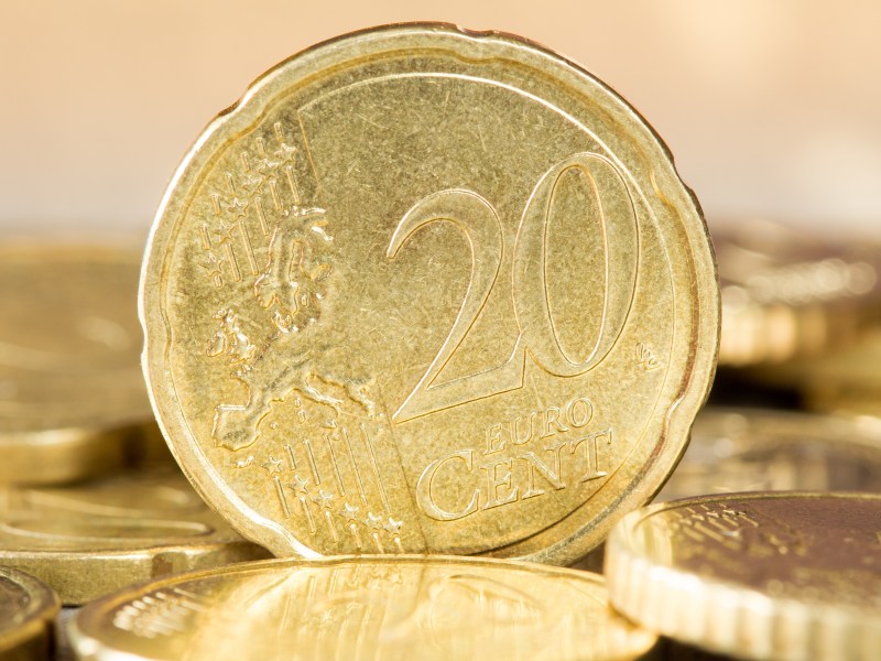 Eine 20-Cent-Euromünze steht senkrecht und mit der Vorderseite nach vorn zwischen weiteren liegenden Cent-Münzen.