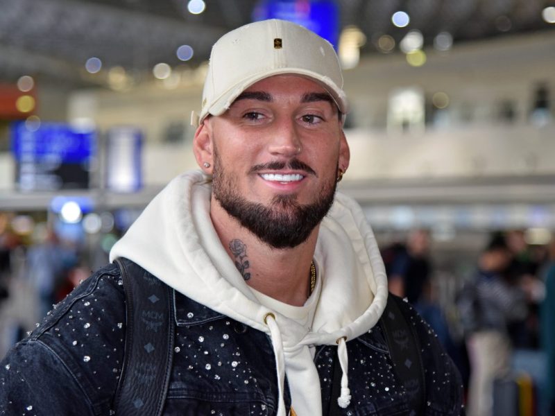 Mike Heiter am Flughafen-terminal. Er trägt eine beige Cap und einen beigen Pullover. Über dem Pullover trägt er eine schwarze Jeansjacke.