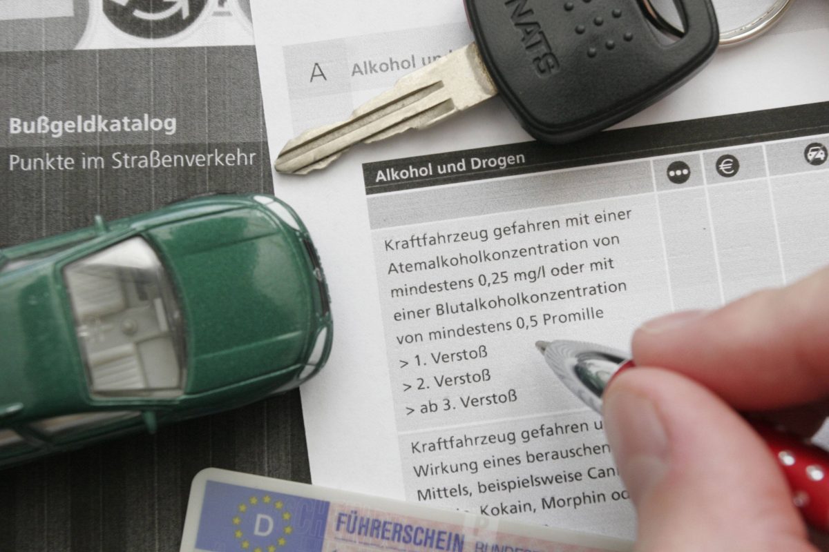 Auf dem Foto sieht man oben rechts einen Autoschlüssel. Unten im Bild ist ein Führerschein abgebildet und links daneben ein grünes Spielzeugauto. Im Hintergrund sind Blätter zu sehen. Links auf dem Blatt steht "Bußgeldkatalog". Auf dem rechten Blatt ist eine Hand mit Kugelschreiber aufgelegt.