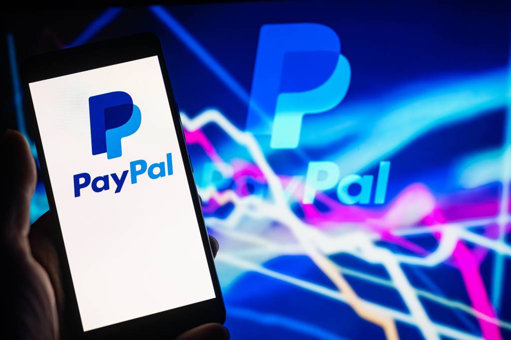 Eine Hand hält ein Smartphone auf dem Paypal geöffnet ist vor einem Hintergrund fallender Aktienkurse.